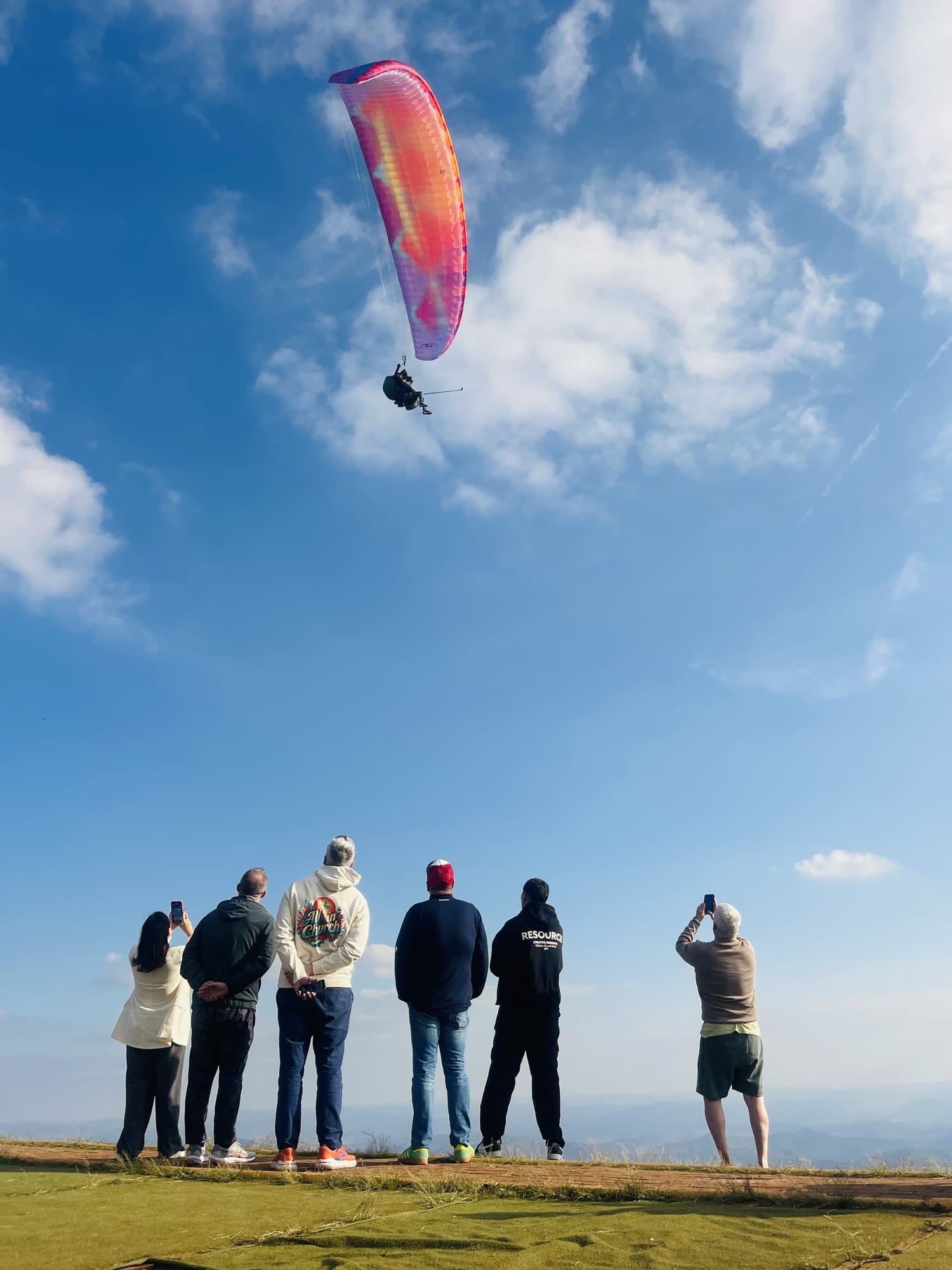 Grupo de pessoas de costas na borda da rampa do Topo do Mundo, na Serra da Moeda, fotografando um parapente que acabou de decolar